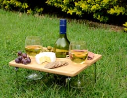 A wooden picnic table with two glasses of white wine, grapes, olives, cheese, and crackers on a grassy surface.