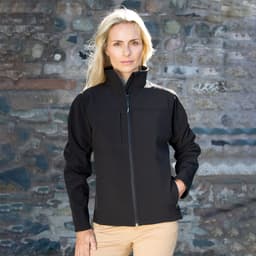A black ladies' classic softshell jacket with a zip front and a chest pocket, worn by a model against a stone wall.