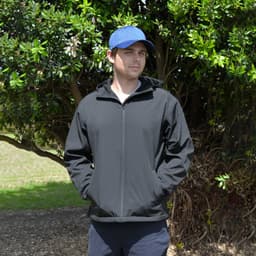A black softshell jacket with a hood, worn by a person standing outdoors among greenery, with a blue cap.