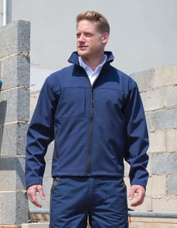 A man wearing a navy soft shell jacket stands against a partially constructed wall, with a branding logo visible.