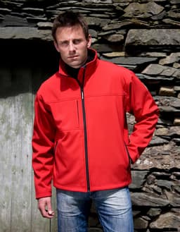 A red soft shell jacket is worn by a model standing against a stone wall, featuring a zip and a slight collar.
