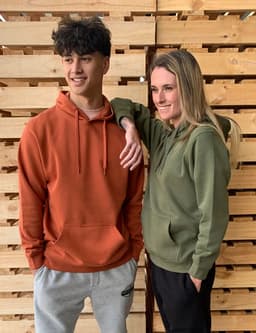 A brown hoodie and a green hoodie are worn by two models against a wooden backdrop. Both hoodies have a front pocket.