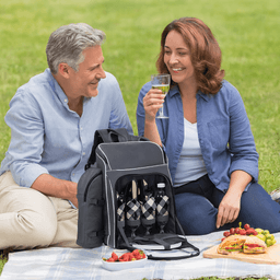 A black picnic backpack with compartments for drink bottles, alongside two people enjoying a picnic on grass.