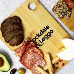 A large bamboo cutting board with assorted food items, including bread, meats, cheese, and an avocado, featuring a logo.