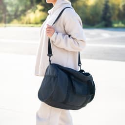 A black RPET duffle bag is being carried by a person wearing a light-coloured hoodie. The bag has an adjustable strap and logo.