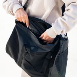 A black RPET duffle bag with multiple compartments, being held by a person. The bag has a shoulder strap and a logo.