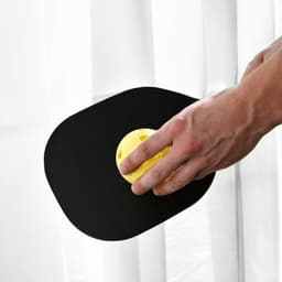 A paddle with a black surface and a yellow pickleball rests against a white curtain.