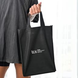A black tote bag with two handles, featuring a logo on one side, held by a person.