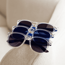 Four pairs of sunglasses sit stacked on a beige surface. The frames are clear, featuring dark blue and black lenses.