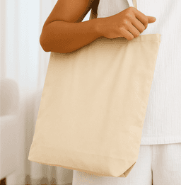 A neutral-toned tote bag held by a person, featuring a simple design and an attached logo.