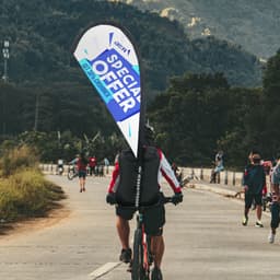 A teardrop flag backpack in blue and white, carried by a cyclist on a path with greenery and mountains in the background.