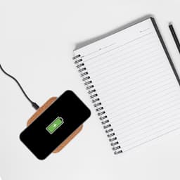 A black smartphone on a square cork wireless charger, placed next to a blank notebook and a pen.
