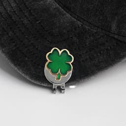 A green and gold soft enamel golf ball marker featuring a four-leaf clover design, resting on a black cap.