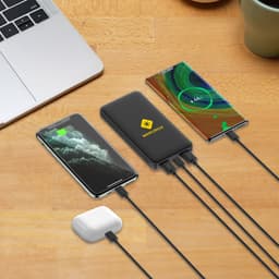A power bank in black with a logo, charging a phone and earbuds on a wooden surface with a laptop and a mug.