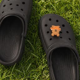 A pair of black clog shoes featuring a 3D bear charm on one shoe, set on green grass.