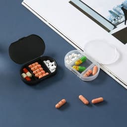 A black pill box with orange and white pills, alongside a clear box containing green and yellow capsules on a dark surface.
