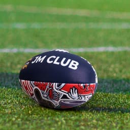 A training AFL ball in dark blue with vibrant red designs, featuring a logo, resting on a grassy field.