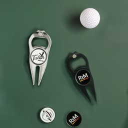 A golf divot tool in silver and a black bottle opener, accompanied by a golf ball and several markers. Both tools feature logos.