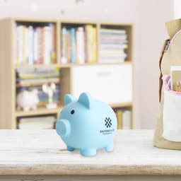 A light blue PVC piggy coin bank with a logo, placed on a wooden table in a room with bookshelves in the background.