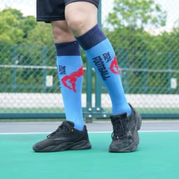 Knee high sports socks in blue with red pattern and a logo, worn with black athletic shoes on a green court.