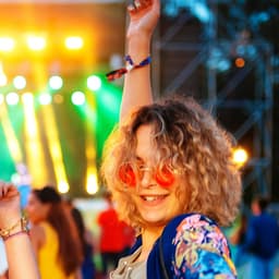 A person with curly hair wearing pink sunglasses and a colourful outfit, raising their arm in a lively outdoor event setting.