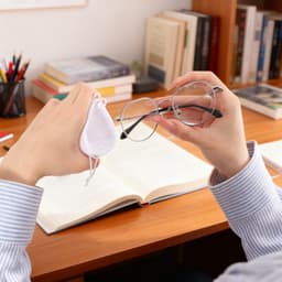 A hand holds glasses and a small white pouch, with books and stationery visible on a wooden desk.