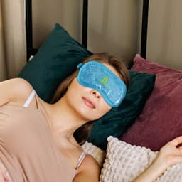 A blue gel cold/hot eye mask rests on a woman's face while she lies on a bed with green and burgundy cushions.