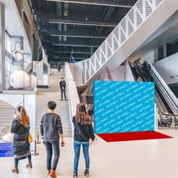 A blue straight fabric popup display with a branding logo, set in a modern atrium. People are walking nearby.