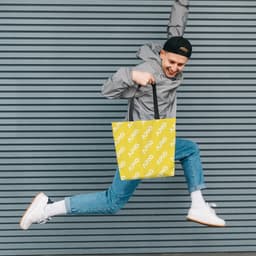 A person jumps while holding a vibrant yellow tote bag featuring a logo, set against a striped grey background.