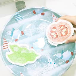 Two colourful, expandable dishwashing sponges in green and pink on a festive patterned plate with bubbles.