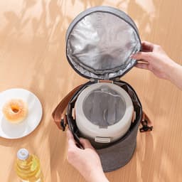 A grey cylindrical lunch bag with a silver interior, being opened to reveal a food container inside. A drink bottle and a donut are nearby.