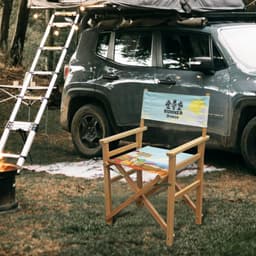 A folding director's chair with a light blue seat featuring a beach design, set near a vehicle in a forested area.