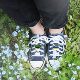 A pair of navy shoes adorned with colourful custom shoe charms, surrounded by small blue flowers and green grass.
