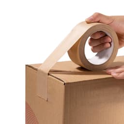 A hand holds a roll of brown self-adhesive kraft paper tape, partially applied to a cardboard box.