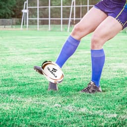 A promotional rugby ball on the ground, featuring a logo, with a player in blue socks preparing to kick it.
