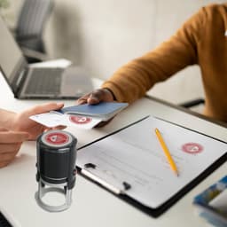 A black custom self-inking stamp is placed on a desk, with hands exchanging a card and paperwork in the background.