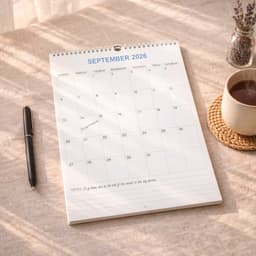 A vertical wall calendar for September 2026, featuring a clean white design, placed on a table with a coffee mug and pen.