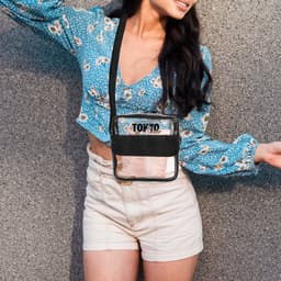 A clear crossbody bag with a black trim, worn over a floral blue top and white shorts.