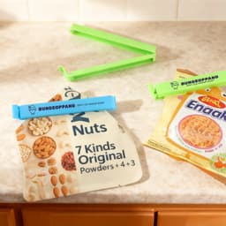 A blue and a green food bag seal clip on packaging, with a variety of nuts and a snack product in the background.