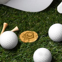 A gold antique golf ball marker placed on grass, surrounded by white golf balls and wooden tees. A golf cap is included.