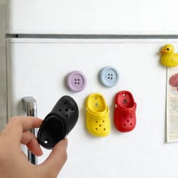A hand holds a black clog shoe fridge magnet, alongside purple, yellow, and red button-shaped magnets on a fridge.