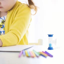 A blue 3-minute sand timer sits beside a child drawing on paper, with colourful pencils scattered in front.