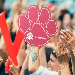 A pink paw foam mitt is raised among a crowd, with hands clapping and cheering in the background.