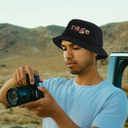 A black Terry towelling bucket hat with a logo, worn by a person holding a camera in a desert landscape.