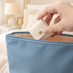 A blue synthetic leather jewellery bag with a hand placing a small cream pouch inside it.
