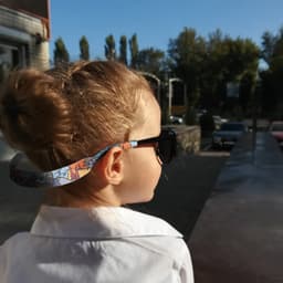 A child wearing black sunglasses with a colourful patterned glasses retainer outdoors, with trees and vehicles in the background.
