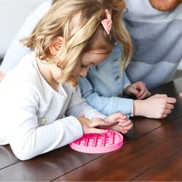 A pink Pop It fidget toy rests on a wooden table, while a child interacts with it, alongside another person.
