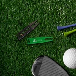 A golf divot tool in black and green, alongside golf tees, a golf ball, and a club, on a grass-like surface.