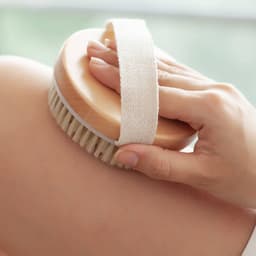 An exfoliating brush with soft massage nodules featuring a wooden base and a fabric strap, being held against skin.