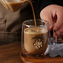 A barrel-shaped double wall mug in clear glass is being filled with a creamy coffee beverage.
